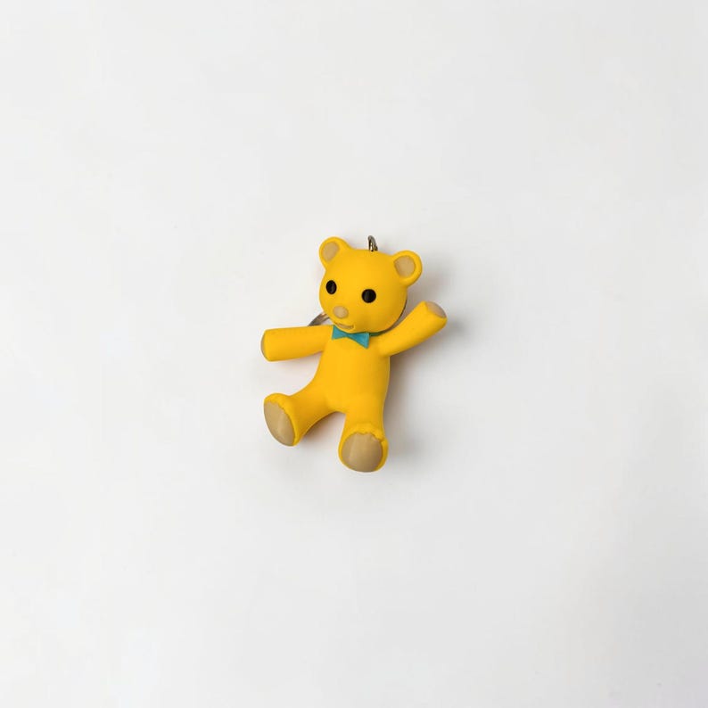 Puede incluir: Un llavero de oso amarillo con una peque&ntilde;a pajarita turquesa en el cuello, ojos negros circulares y detalles en color canela claro en las orejas, manos y pies. El oso aparece en posici&oacute;n sentada con un brazo levantado y una anilla met&aacute;lica.
