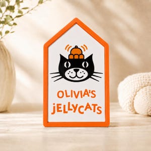 以下が含まれることがあります： オレンジ色のフレームの看板で、白い内側に帽子をかぶった黒猫の顔とオレンジ色の「OLIVIA'S JELLYCATS」の文字が描かれています。 看板は家の形をしています。