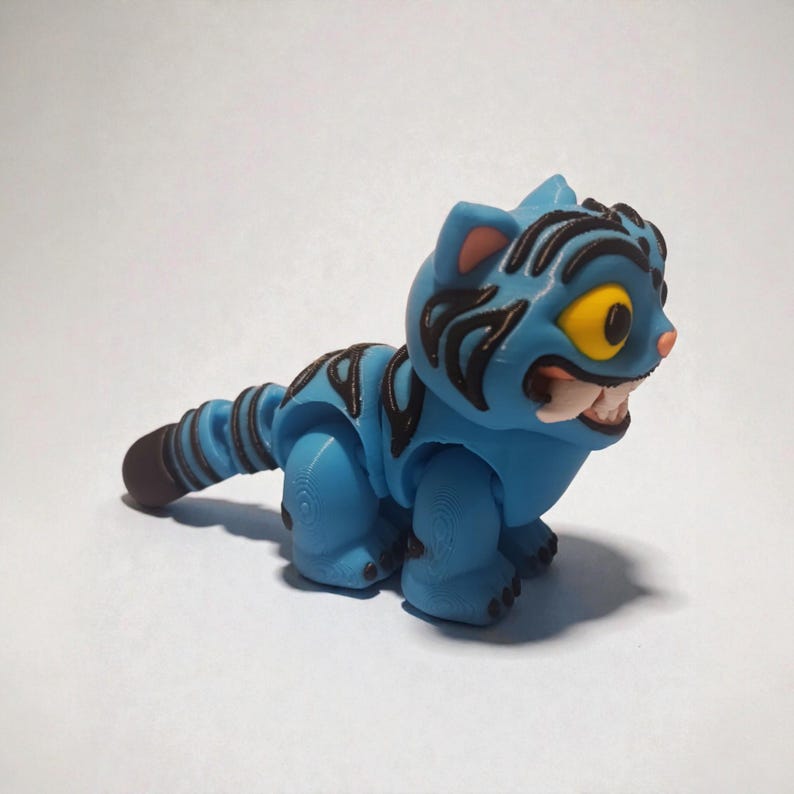 Puede incluir: Un juguete de gato azul con rayas negras y ojos amarillos. El gato tiene una amplia sonrisa, dientes visibles y una cola marr&oacute;n. El juguete est&aacute; hecho de pl&aacute;stico y tiene un dise&ntilde;o de dibujos animados.