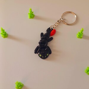 Op de afbeelding: Een zwarte draak sleutelhanger met groene ogen en een rode accent, bevestigd aan een goudkleurige sleutelring. Verschillende kleine, groene, boomvormige beeldjes zijn rond de sleutelhanger verspreid.