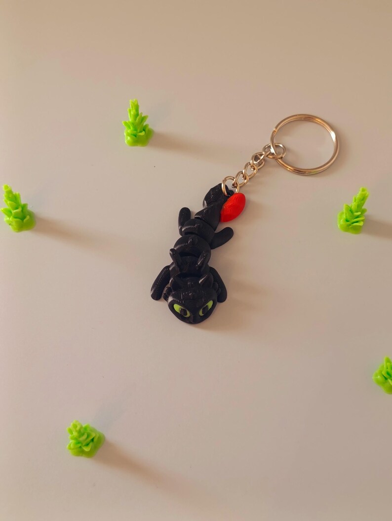 Op de afbeelding: Een zwarte draak sleutelhanger met groene ogen en een rode accent, bevestigd aan een goudkleurige sleutelring. Verschillende kleine, groene, boomvormige beeldjes zijn rond de sleutelhanger verspreid.