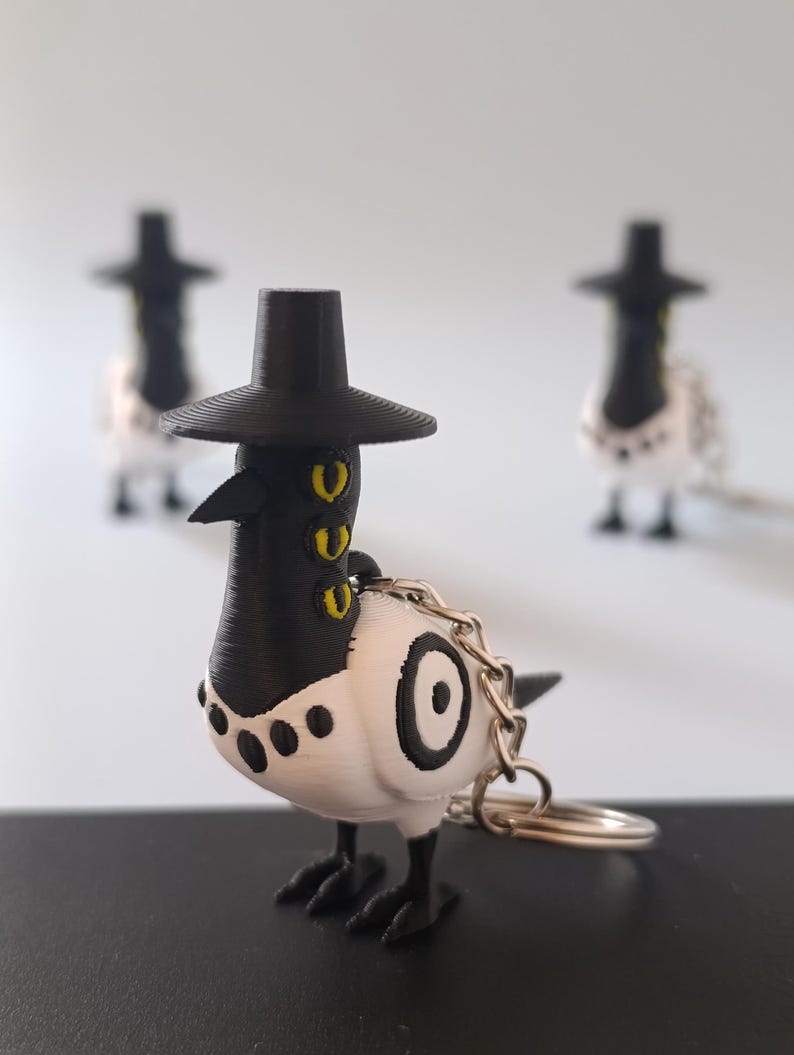 May include: A 3D-printed keychain featuring a white and black bird with a black hat. The bird has yellow eyes and a circular design on its side. A silver keyring is attached. Two similar, blurred figures are in the background.