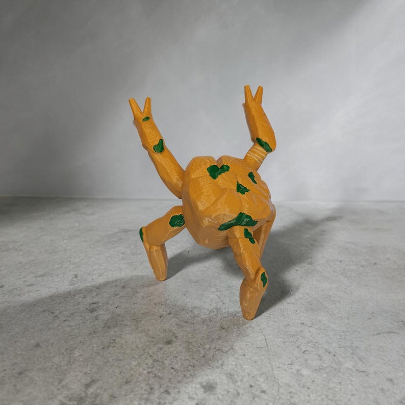 May include: A wooden, abstract figurine in shades of orange and green. The figurine has a rounded body with four legs and two arms raised in a V-shape. Green paint accents the body and limbs. The figurine is set against a neutral gray background.