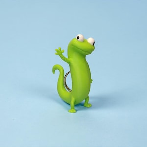 May include: A bright green gecko keychain with a friendly wave. The gecko has large white eyes and a curled tail. The keychain is made of a smooth, glossy material and is set against a light blue background.