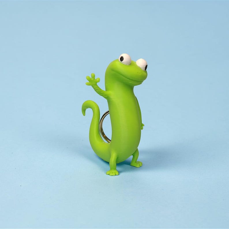 May include: A bright green gecko keychain with a friendly wave. The gecko has large white eyes and a curled tail. The keychain is made of a smooth, glossy material and is set against a light blue background.
