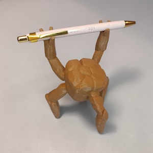 Pode incluir: Uma figura articulada castanha segura uma caneta branca com detalhes dourados. A caneta tem texto impresso. A figura tem um design texturizado e geom&eacute;trico e est&aacute; posada como se estivesse a levantar a caneta.