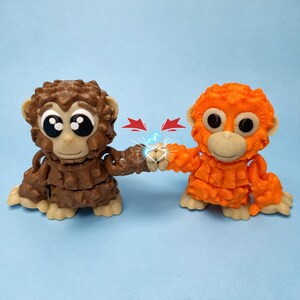 May include: Two toy orangutans, one brown and one orange, are fist-bumping. The brown orangutan is on the left, and the orange one is on the right. Both have large, expressive eyes and are in a playful pose. Red arrows and blue lightning bolts are between their fists.