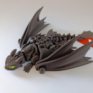K&ouml;nnte beinhalten: Dunkelbraune Drachenfigur mit beweglichen Fl&uuml;geln und Gliedma&szlig;en. Der Drache hat gr&uuml;ne Augen und einen detaillierten, strukturierten K&ouml;rper. Die Fl&uuml;gel sind ausgebreitet, und der Drache liegt. Die Figur ist wahrscheinlich ein Sammlerst&uuml;ck oder ein Spielzeug.