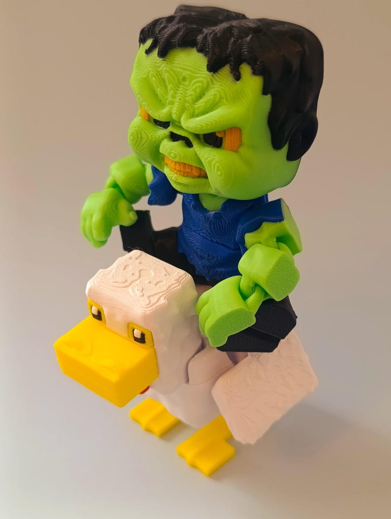 May include: A 3D-printed figurine of a green-skinned monster with black hair and a blue shirt riding a white and yellow duck. The monster has an angry expression. The duck has a yellow beak and feet.