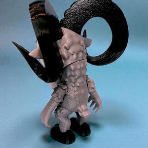 May include: A gray and black figurine of a ram-like creature with large curved horns. The figurine has a textured body and is posed with its back to the viewer. The horns are black, and the body is gray. The figurine is on a light blue surface.