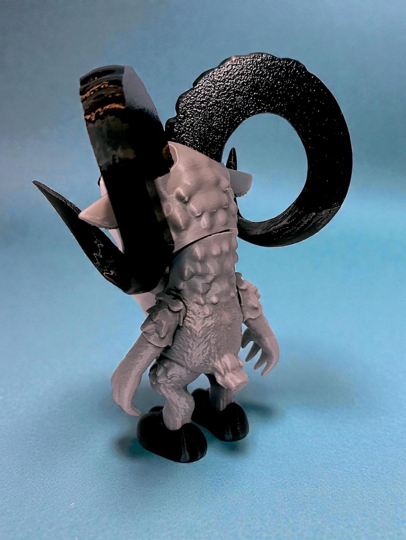 May include: A gray and black figurine of a ram-like creature with large curved horns. The figurine has a textured body and is posed with its back to the viewer. The horns are black, and the body is gray. The figurine is on a light blue surface.