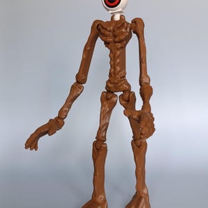May include: A brown articulated figure with a large eye for a head. The eye is white with a red and black iris. The figure has a skeletal appearance with jointed limbs and feet. The figure is standing against a light gray background.