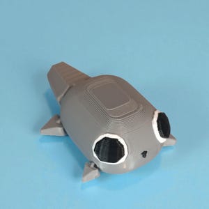 May include: A gray 3D-printed toy shaped like a stylized fish or aquatic creature. It has black eyes with white outlines, small fins, and a textured surface. The toy is set against a light blue background.