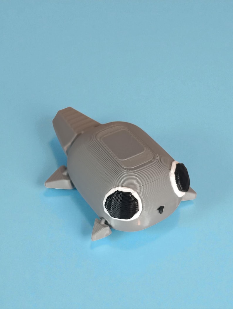 May include: A gray 3D-printed toy shaped like a stylized fish or aquatic creature. It has black eyes with white outlines, small fins, and a textured surface. The toy is set against a light blue background.