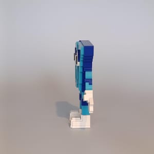 Pode incluir: Uma figura pixelizada em tons de azul, branco e preto, constru&iacute;da a partir de pequenos blocos. A figura tem uma cabe&ccedil;a retangular, um corpo com tons variados de azul e p&eacute;s brancos. A figura est&aacute; em p&eacute;.