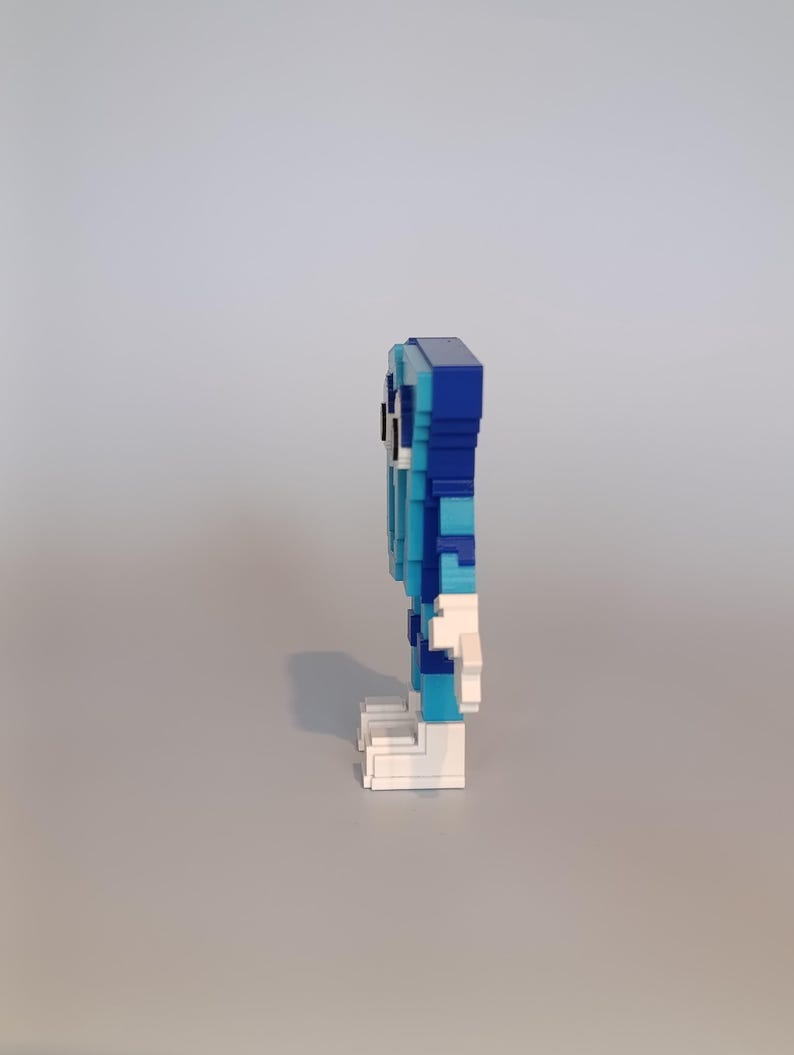Pode incluir: Uma figura pixelizada em tons de azul, branco e preto, constru&iacute;da a partir de pequenos blocos. A figura tem uma cabe&ccedil;a retangular, um corpo com tons variados de azul e p&eacute;s brancos. A figura est&aacute; em p&eacute;.