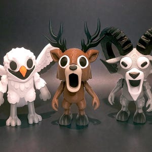 May include: Three whimsical animal figurines: an owl with outstretched wings, a deer with antlers, and a ram with large horns. The owl is white with an orange beak, the deer is brown, and the ram is gray.