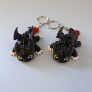 May include: Two black dragon keychains with green eyes, white paws, and red accents. The dragon figures have detailed wings and are attached to silver key rings. The keychains are lying on a light blue surface.