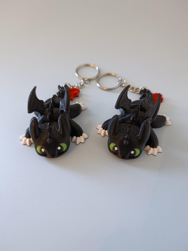 May include: Two black dragon keychains with green eyes, white paws, and red accents. The dragon figures have detailed wings and are attached to silver key rings. The keychains are lying on a light blue surface.