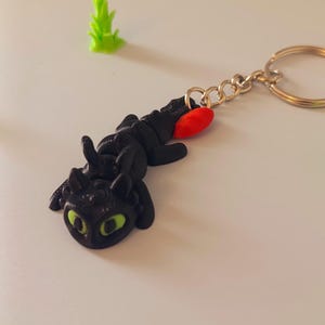 Op de afbeelding: Zwarte draken sleutelhanger met groene ogen en een rode accent, bevestigd aan een goudkleurige sleutelring. Er is ook een kleine, groene, plantachtige figuur aanwezig. De draak ligt, en de sleutelhanger is waarschijnlijk een verzamelobject of accessoire.