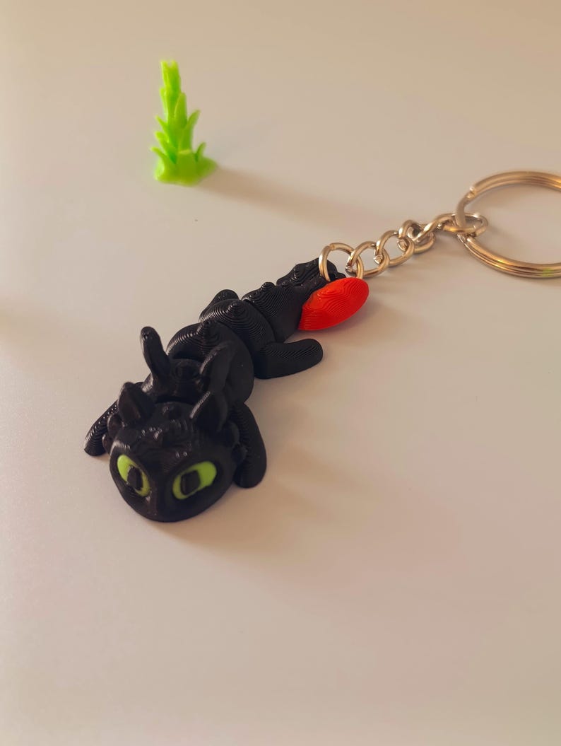 Op de afbeelding: Zwarte draken sleutelhanger met groene ogen en een rode accent, bevestigd aan een goudkleurige sleutelring. Er is ook een kleine, groene, plantachtige figuur aanwezig. De draak ligt, en de sleutelhanger is waarschijnlijk een verzamelobject of accessoire.