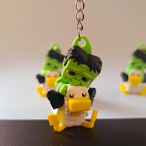 Op de afbeelding: Een groene en zwarte Hulk-achtige figuur rijdt op een witte en gele eenden sleutelhanger. De sleutelhanger heeft een gouden ketting en een groene lus. Extra vergelijkbare sleutelhangers zijn zichtbaar op de achtergrond.