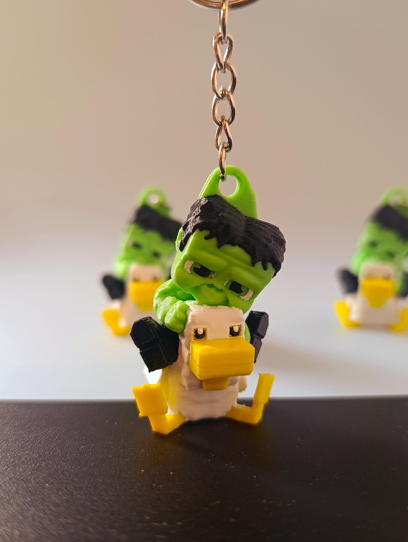 Op de afbeelding: Een groene en zwarte Hulk-achtige figuur rijdt op een witte en gele eenden sleutelhanger. De sleutelhanger heeft een gouden ketting en een groene lus. Extra vergelijkbare sleutelhangers zijn zichtbaar op de achtergrond.