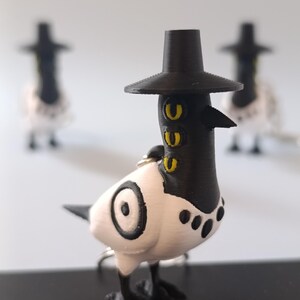 May include: A 3D-printed bird figurine with a black hat, beak, and feet, and a white body with black accents. The bird has yellow eye details and a keychain ring. Two other similar figurines are blurred in the background.