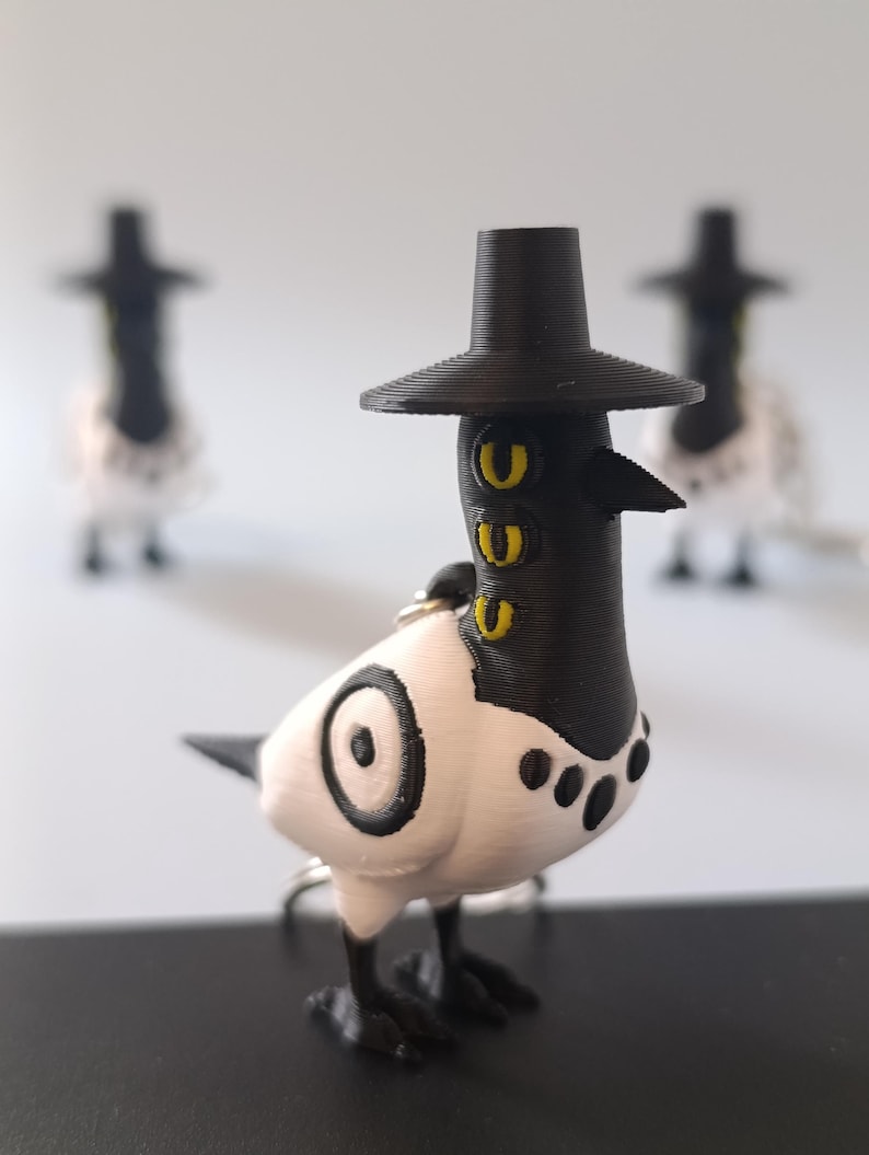 May include: A 3D-printed bird figurine with a black hat, beak, and feet, and a white body with black accents. The bird has yellow eye details and a keychain ring. Two other similar figurines are blurred in the background.