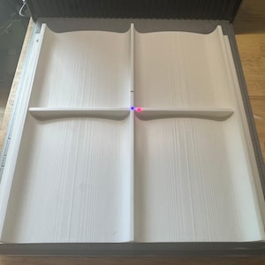 May include: A white wooden book-shaped object with a shelf. The object is made of light-coloured wood and has a textured surface. The object is in a grey frame and is on a wooden surface.