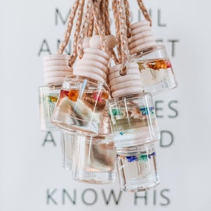 May include: A collection of small, clear glass bottles filled with liquid and dried flowers, suspended by braided cords. Each bottle has a wooden cap. The background features the words "SO GO AND KNOW HIS NAME".