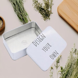 Op de afbeelding: Een rechthoekig wit metalen blik met een afneembaar deksel. Op het deksel staat de zwarte tekst "DESIGN YOUR OWN" in een eenvoudig schreefloos lettertype. De binnenkant van het blik is metaalzilver en bevat een klein rond logo op de bodem.