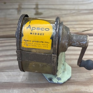 May include: Vintage Apsco Midget pencil sharpener with a yellow label. The label includes the text "Apsco Midget" and "Apsco products inc. Beverly Hills, Calif. Rockford, Ill., U.S.A." The sharpener has a green base and a metal handle.