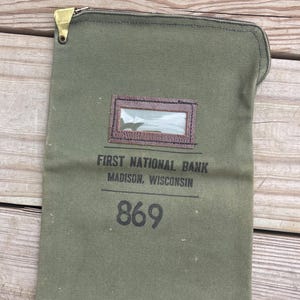 May include: An olive green canvas bank deposit bag with a metal zipper closure. It features a brown leather-trimmed clear plastic ID window and black printed text reading FIRST NATIONAL BANK MADISON, WISCONSIN above the large black number 869.
