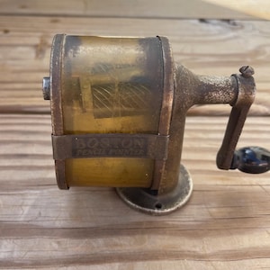 May include: An antique Boston Pencil Pointer pencil sharpener. The sharpener is made of metal with a clear, yellowed plastic window. The words "BOSTON PENCIL POINTER" are printed on a metal band. The handle has a black knob.