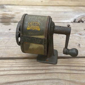 May include: Vintage Giant pencil sharpener made by Automatic Pencil Sharpener Co. Chicago, USA. The sharpener is made of metal and has a hand crank. The body is olive green with a yellow and black label.