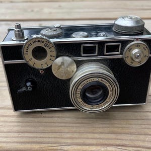 May include: Vintage black and silver camera with multiple dials and a lens. The camera has a range finder and a textured black body. The lens is marked "COATED CINTAR ARGUS". The camera is on a wooden surface.
