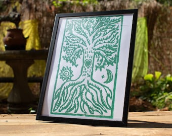 Puede incluir: Una impresión artística enmarcada con un diseño de árbol verde que incluye un corazón, un sol y una luna. La obra de arte está sobre un fondo blanco y enmarcada en un marco negro. La impresión se muestra sobre una superficie de madera.