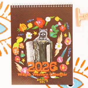May include: A spiral-bound 2026 calendar with a brown cover featuring a raccoon playing an accordion, surrounded by colorful flowers. The text "Wild & Western Calendar" and "Stationery Bakery" are also visible.