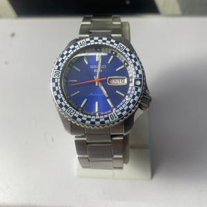 Puede incluir: Un reloj Seiko de acero inoxidable con esfera azul y bisel a cuadros azul y blanco. El reloj tiene una pantalla de fecha que muestra "DOM 19" y un segundero rojo. La correa del reloj también es de acero inoxidable.