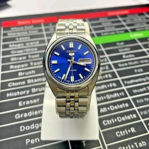 May include: A stainless steel Seiko automatic wristwatch with a blue dial. The watch has a silver band and a day-date display. The watch is displayed on a white stand.