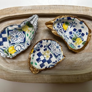 Op de afbeelding: Drie decoratieve oesterschelpen met blauw-witte tegelpatronen, gele citroenen en bloemaccenten. Eén schelp heeft een zilveren rand en twee hebben gouden randen. Ze liggen op een houten dienblad.