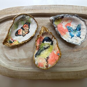 Puede incluir: Tres conchas de ostra decorativas con diseños pintados. Una concha presenta una mariposa naranja, otra una mariposa azul y la tercera un pájaro pequeño. Las conchas tienen bordes metálicos dorados y plateados y se exhiben en una bandeja de madera.