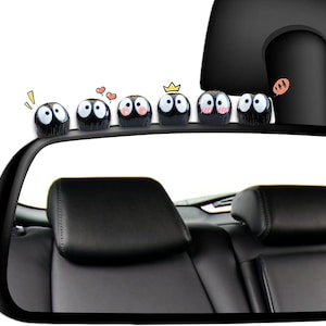 May include: A collection of six black, cartoon-like figures with large white eyes and various facial expressions, perched atop a car's rearview mirror. Each figure has a unique design, including hearts, a crown, and a speech bubble.