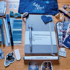 May include: A gray A5 ruled notebook with a silver heart-topped pen, a blue pencil case with Let it Snow text, various blue-toned pens, markers, stickers, sticky tabs, a white keychain, and a candy cane-striped pen on a wooden surface.
