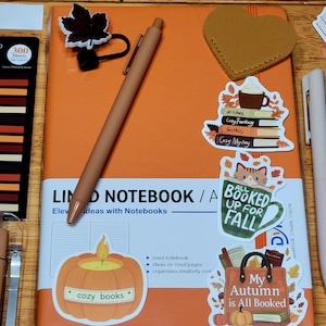 May include: An orange notebook with the text "LIND NOTEBOOK / A" and stickers with fall themes. A brown pen and a yellow Sharpie marker are also visible. The stickers include a pumpkin, a coffee cup, and the text "My Autumn is All Booked."
