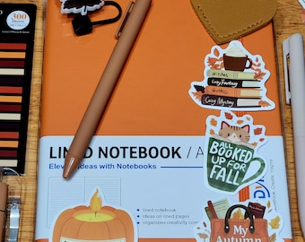 May include: An orange notebook with the text "LIND NOTEBOOK / A" and stickers with fall themes. A brown pen and a yellow Sharpie marker are also visible. The stickers include a pumpkin, a coffee cup, and the text "My Autumn is All Booked."