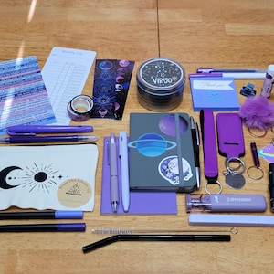 May include: A collection of stationery and accessories in shades of purple, including pens, a notebook, stickers, a keychain, and a small tin. The items are arranged on a wooden surface, creating a visually appealing flat lay.