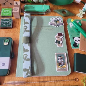 May include: An assortment of stationery items in shades of green and brown, including stamps, washi tape, a notebook with the text "WRITE IT DOWN", and stickers. A green A7 notebook with a cactus charm is also visible.