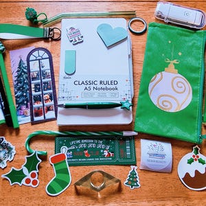 May include: A collection of stationery items including a white A5 notebook, a green pencil case with a gold ornament print, green pens, a candy cane-shaped pen, stickers, bookmarks, and a small white electronic device on a wooden surface.